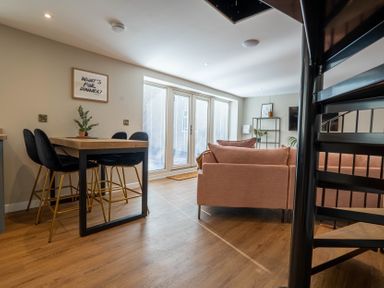 A living room and dining area with a wooden table and black chairs next to pink sofas and glass doors at Coral Cottage in Kessingland