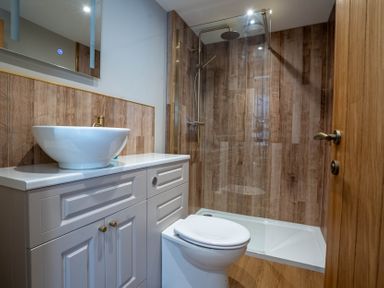 A bathroom with a white sink on a gray cabinet a toilet and a glass shower with wooden wall panels at Coral Cottage in Kessingland