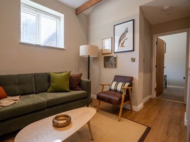 A living room with a green sofa pillows and a floor lamp next to a brown armchair with framed pictures on the wall at Dunes Cottage in Kessingland