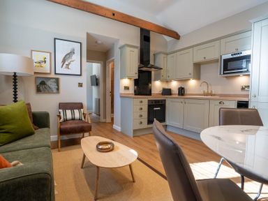 A living room with green sofa brown chair and a wooden coffee table next to a kitchen with light cabinets and a dining table at Dunes Cottage in Kessingland