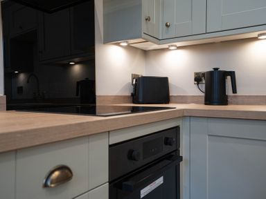 A kitchen corner with a black toaster and kettle on a wooden countertop above built in oven and light grey cabinets at Dunes Cottage in Kessingland