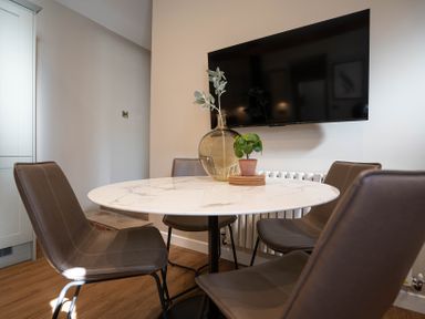 A dining area with a round marble table, four brown chairs, two plants on the table, and a wall-mounted television at Dunes Cottage in Kessingland
