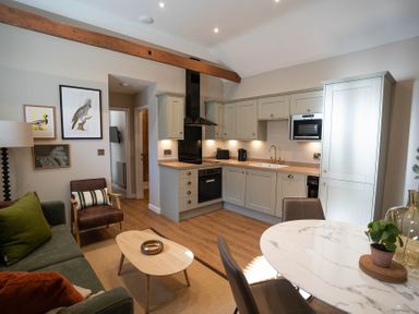 A kitchen and living area with cabinets, a sofa, armchair, coffee table, dining table, and framed pictures at Dunes Cottage in Kessingland