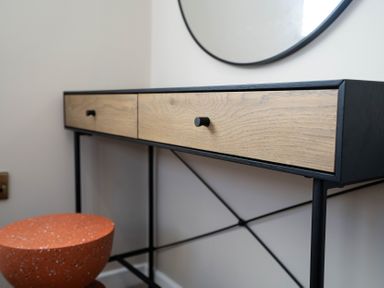 A wooden drawer console table with black frame and a round mirror above it and an orange stool below at Dunes Cottage in Kessingland