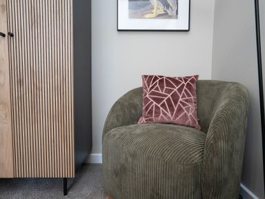 A corner with a green upholstered armchair with a patterned pillow next to a wooden cabinet with vertical grooves in a room at Dunes Cottage in Kessingland