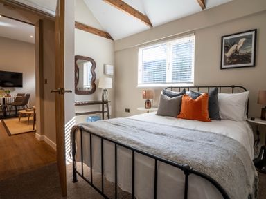 A bedroom with a bed pillows and bedside tables next to a window and an open door showing a dining area at Dunes Cottage in Kessingland