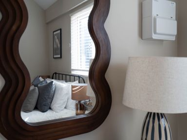 A reflection in a wavy wooden mirror showing a bed with pillows a table lamp window blinds and a framed picture at Dunes Cottage in Kessingland