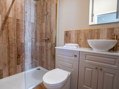 A bathroom with a shower area with wooden paneling a toilet a countertop with a vessel sink and folded towels at Dunes Cottage in Kessingland