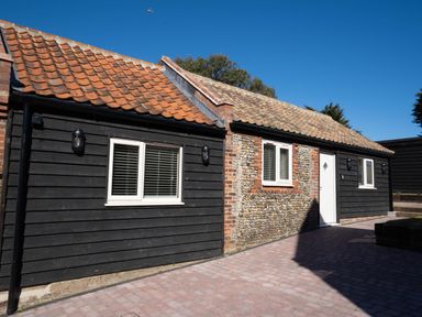 An exterior view of a single story building with black wood siding and pebble stone walls with white windows and a white door at Dunes Cottage in Kessingland