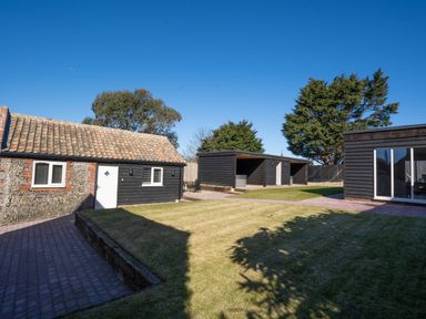 A backyard with a lawn, a brick path, a small building with tiled roof, and black wooden structures at Dunes Cottage in Kessingland
