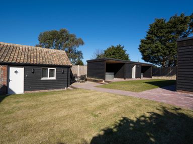 An outdoor area with black wooden buildings a grassy lawn and a paved walkway at Dunes Cottage in Kessingland