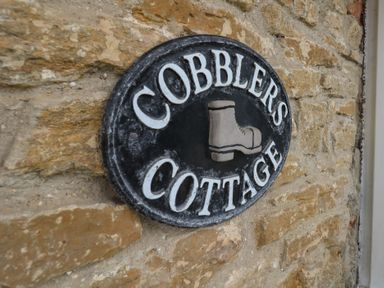 A round black sign with white letters reading Cobblers Cottage and a boot image on a stone wall at Cobblers Cottage in Langton Herring