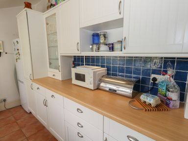 A kitchen countertop with a microwave a bread bin and food items on a wooden counter with white cabinets and blue tiled backsplash at Cobblers Cottage in Langton Herring