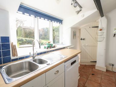 A kitchen sink under a window with a blue blind a wooden countertop a white dishwasher a hanging wire basket and a wooden door at Cobblers Cottage in Langton Herring