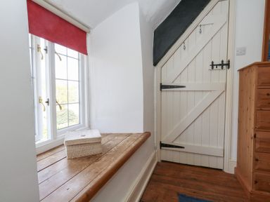A window with a red blind and a wicker basket on a wooden sill next to a white wooden door with black hinges and a wooden chest of drawers at Cobblers Cottage in Langton Herring