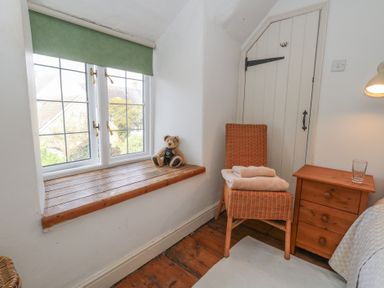 A corner of a bedroom with a wooden window seat with a teddy bear a wicker chair with folded towels a wooden nightstand with a glass and a white door at Cobblers Cottage in Langton Herring
