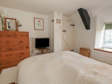 A bedroom with a bed covered in a white textured blanket a wooden dresser with a mirror a small television on a stand a wicker basket a window with a green blind and a white door with black hinges at Cobblers Cottage in Langton Herring