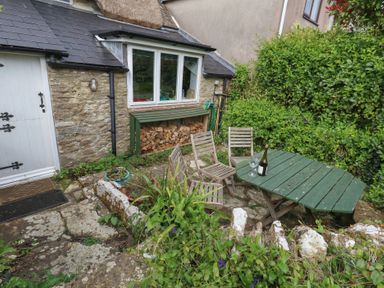 An outdoor patio area with a green wooden table and chairs beside a stone wall and a house with stacked firewood at Cobblers Cottage in Langton Herring