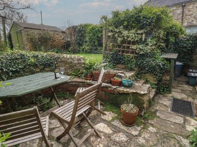An outdoor patio with wooden chairs and a table with drinks and potted plants around at Cobblers Cottage in Langton Herring