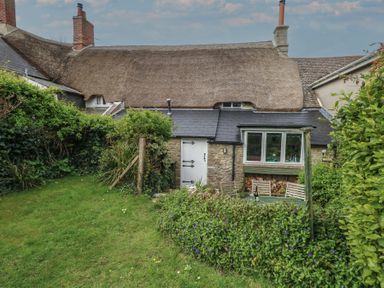 A garden with green grass and plants in front of a stone cottage with a thatched roof and wooden chairs at Cobblers Cottage in Langton Herring