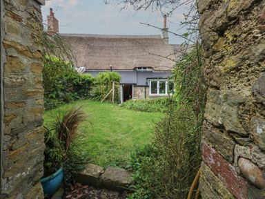 A grassy backyard garden area with plants and stone walls leading to a thatched roof cottage at Cobblers Cottage in Langton Herring