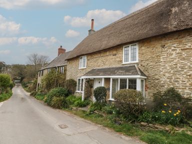 A stone cottage with a thatched roof and garden by a rural road at Cobblers Cottage in Langton Herring