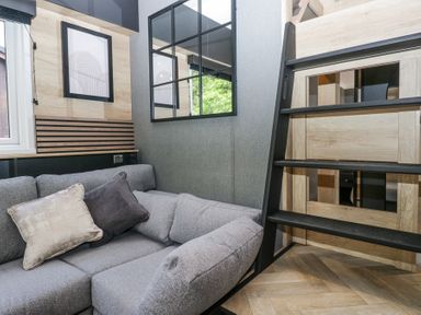 A corner of a room with a gray sofa with cushions a window a black framed wall mirror and a wooden ladder at Lingy Luxury Family Pod in Keswick