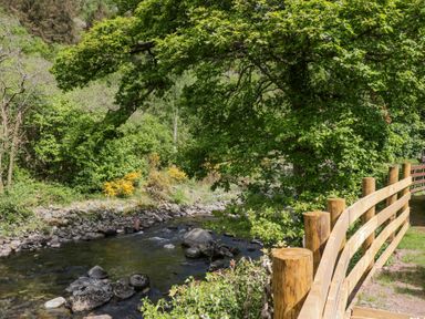 A river flowing beside a wooden fence with trees and bushes in the background at Lingy Luxury Family Pod in Keswick