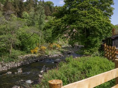 A river flowing through a wooded area with a wooden fence and a building on the right at Lingy Luxury Family Pod in Keswick