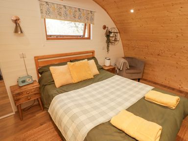 A bedroom with a double bed green bedding and yellow pillows beside a small wooden nightstand and a gray chair at Cabanau Bach in Bedwas