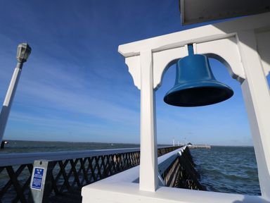 A blue bell hanging on a white wooden structure by a long pier extending into the sea at Plimoth N9 in Yarmouth