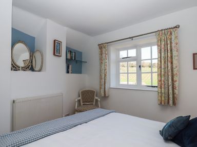 A bedroom with a window with floral curtains an armchair a wall shelf with books and a mirror at Badger's Den in Budleigh Salterton