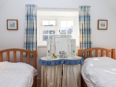 A bedroom with two single wooden beds and a vanity table under a window with blue and white curtains at Badger's Den in Budleigh Salterton
