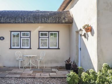 An outdoor patio with a small white table and two chairs in front of a cottage with two windows and a plant on the wall at Badger's Den in Budleigh Salterton