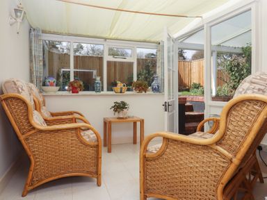 A sunroom with wicker chairs a wooden table with a plant and glass doors leading to a garden at Badger's Den in Budleigh Salterton