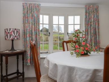 A dining room with floral curtains round table with flower centerpiece and a side table with lamp at Badger's Den in Budleigh Salterton