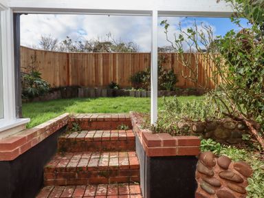 A small patio with brick steps leading up to a grassy garden enclosed by a wooden fence with plants along the edges at Badger's Den in Budleigh Salterton