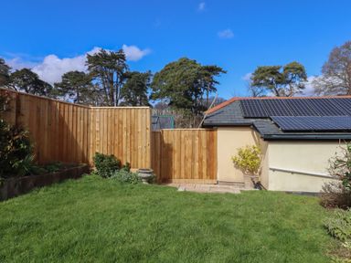 A backyard with a grassy lawn wooden fencing and a building with solar panels on the roof at Badgers Den in Budleigh Salterton