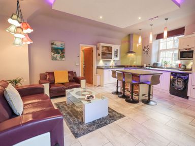 An open living room and kitchen with two sofas a glass coffee table a kitchen island with four stools and kitchen appliances at 1 The Old Coach House in Ambleside