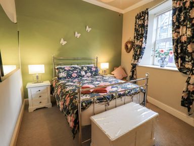A bedroom with a metal bed frame floral bedding two bedside tables with lamps a window with floral curtains and a storage chest at 1 The Old Coach House in Ambleside