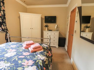 A bedroom with a floral bedspread and folded towels on the bed a white wardrobe and dresser with a vase and a TV mounted on the wall at 1 The Old Coach House Ambleside