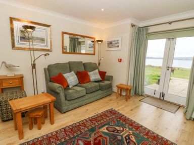 A sitting room with green sofa red and check cushions wooden tables floor lamps and glass doors opening to a garden at Carningli Suite near Newport