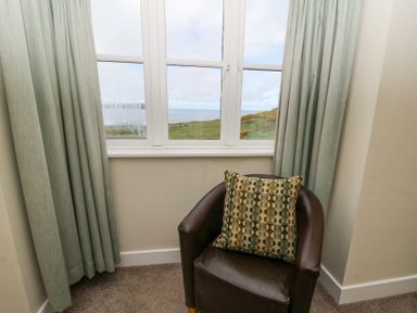 A brown armchair with a patterned cushion in front of a window with green curtains and a view of hills and the sea at the Carningli Suite near Newport