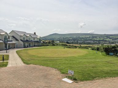 A golf tee area with a sign indicating 10th tee and houses beside it near a grassy landscape and hills in the background at Carningli Suite near Newport