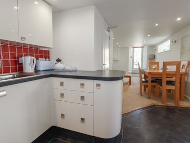 A kitchen area with white cabinets red tiled backsplash and black countertop next to a dining area with wooden table and chairs at St Georges in Padstow