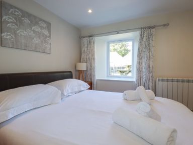 A bedroom with a double bed white linens and rolled towels on the bed a window with curtains a bedside table with a lamp and a painting on the wall at St Georges in Padstow