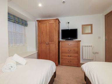 A bedroom with two beds a wooden wardrobe and dresser a small television and a window with a blue and green valance at St Georges in Padstow