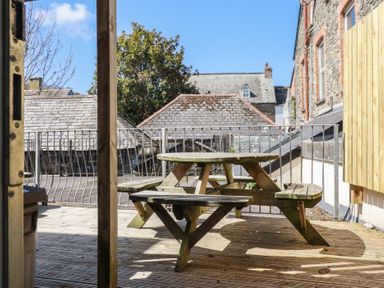 A wooden picnic table on a deck with metal railing and stone buildings in the background at St Georges in Padstow