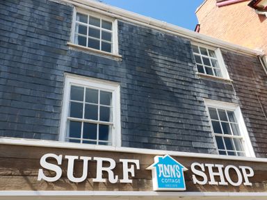 The exterior of a building with slate tiles and a wooden sign that reads surf shop with a blue sign for anns cottage