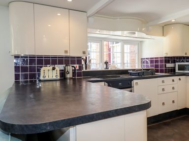 A kitchen with dark countertops white cabinets purple tiled backsplash and appliances at St Saviours in Padstow